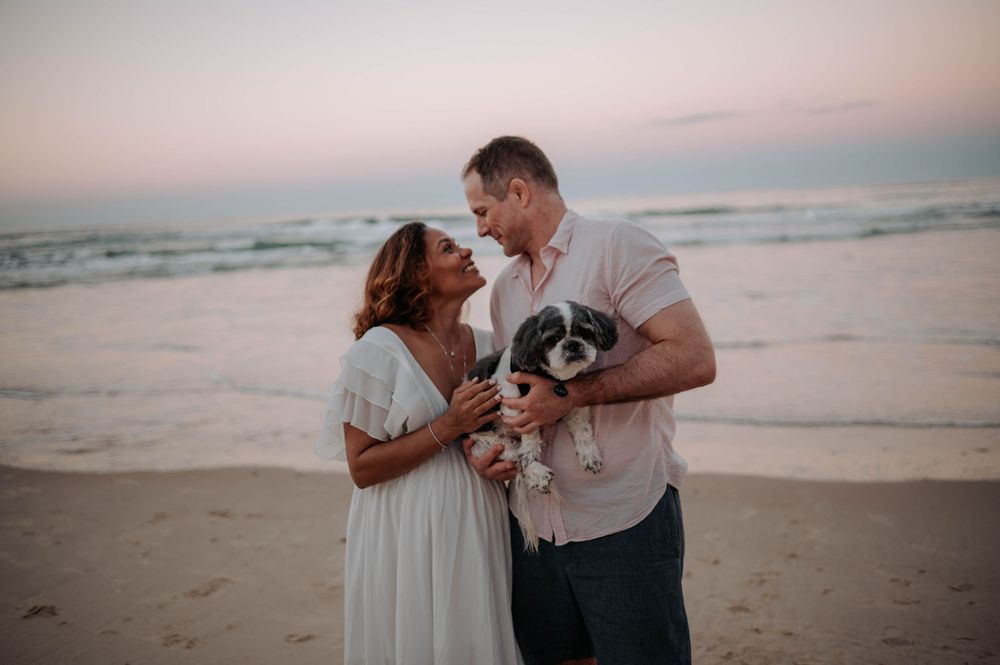 Couple stands on beach at sunset holding their small black and white dog while sharing a romantic moment.
