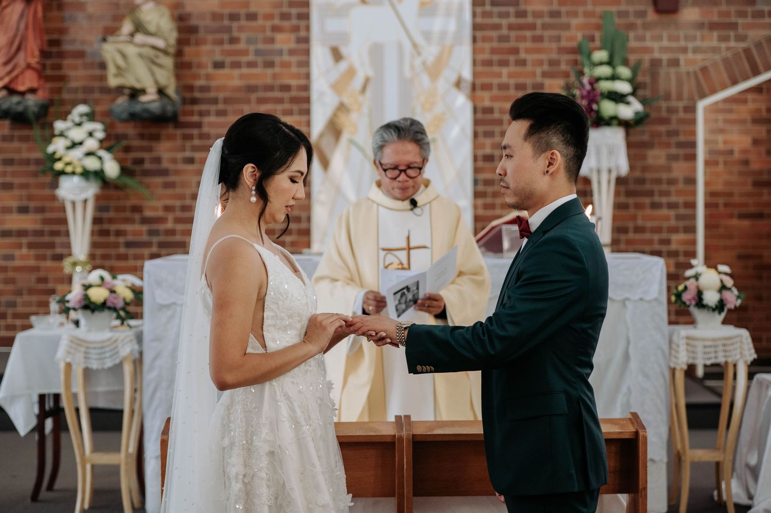 Wedding ceremony takes place in a brick church with white floral arrangements and a priest presiding at the altar.