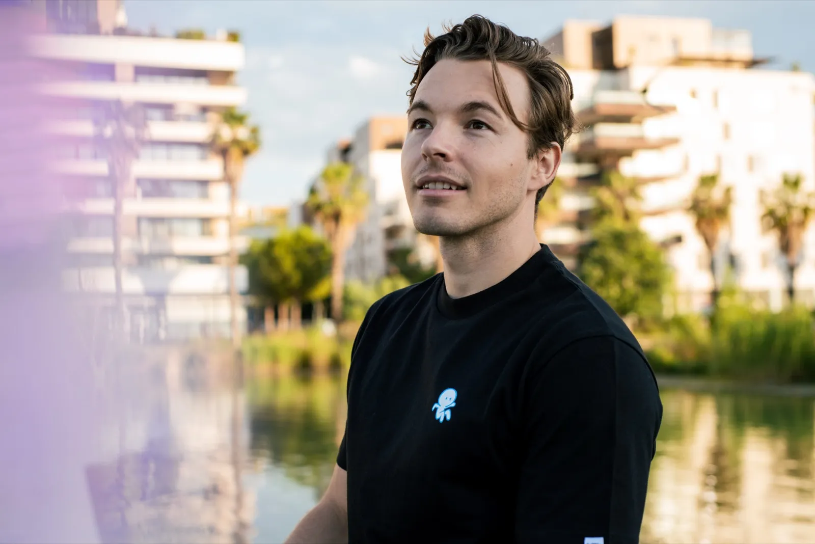 Portrait d’un homme souriant en t-shirt noir, debout au bord de l’eau avec des immeubles et des palmiers en arrière-plan.
