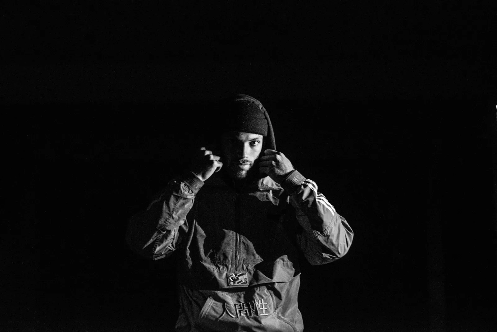 Homme en tenue sombre, capuche sur la tête, dans une ambiance sombre et contrastée, gestuelle expressive.