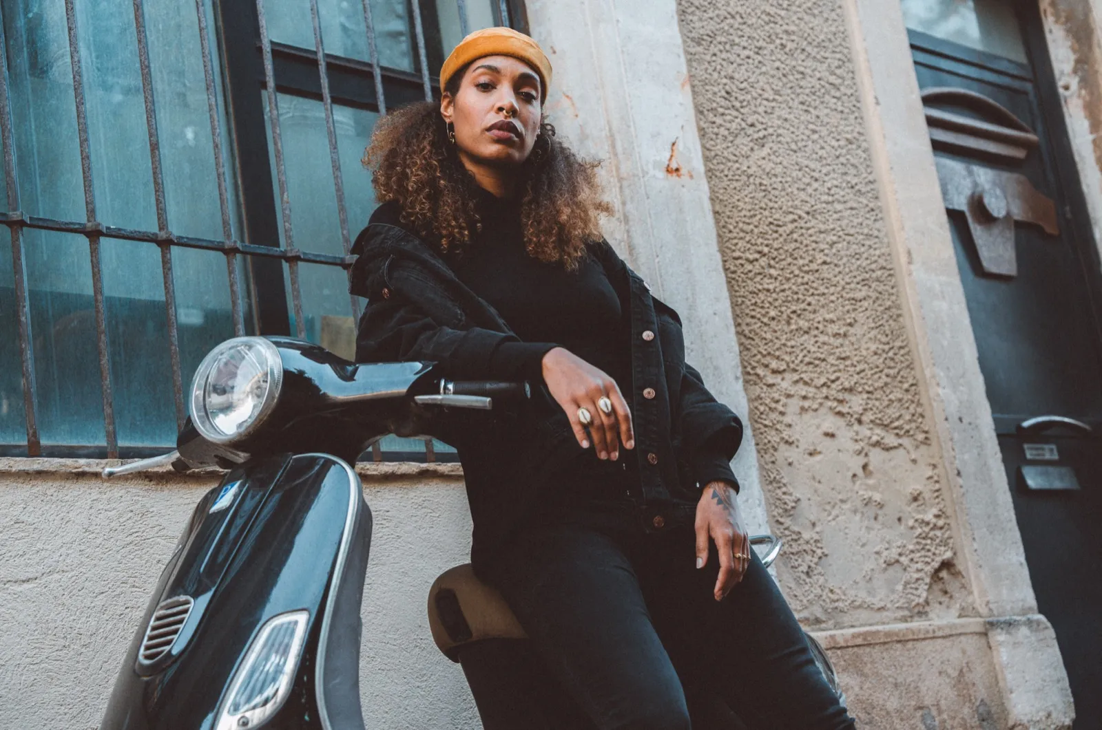 Femme assise sur un scooter devant un mur beige, regard confiant et bonnet sur la tête.