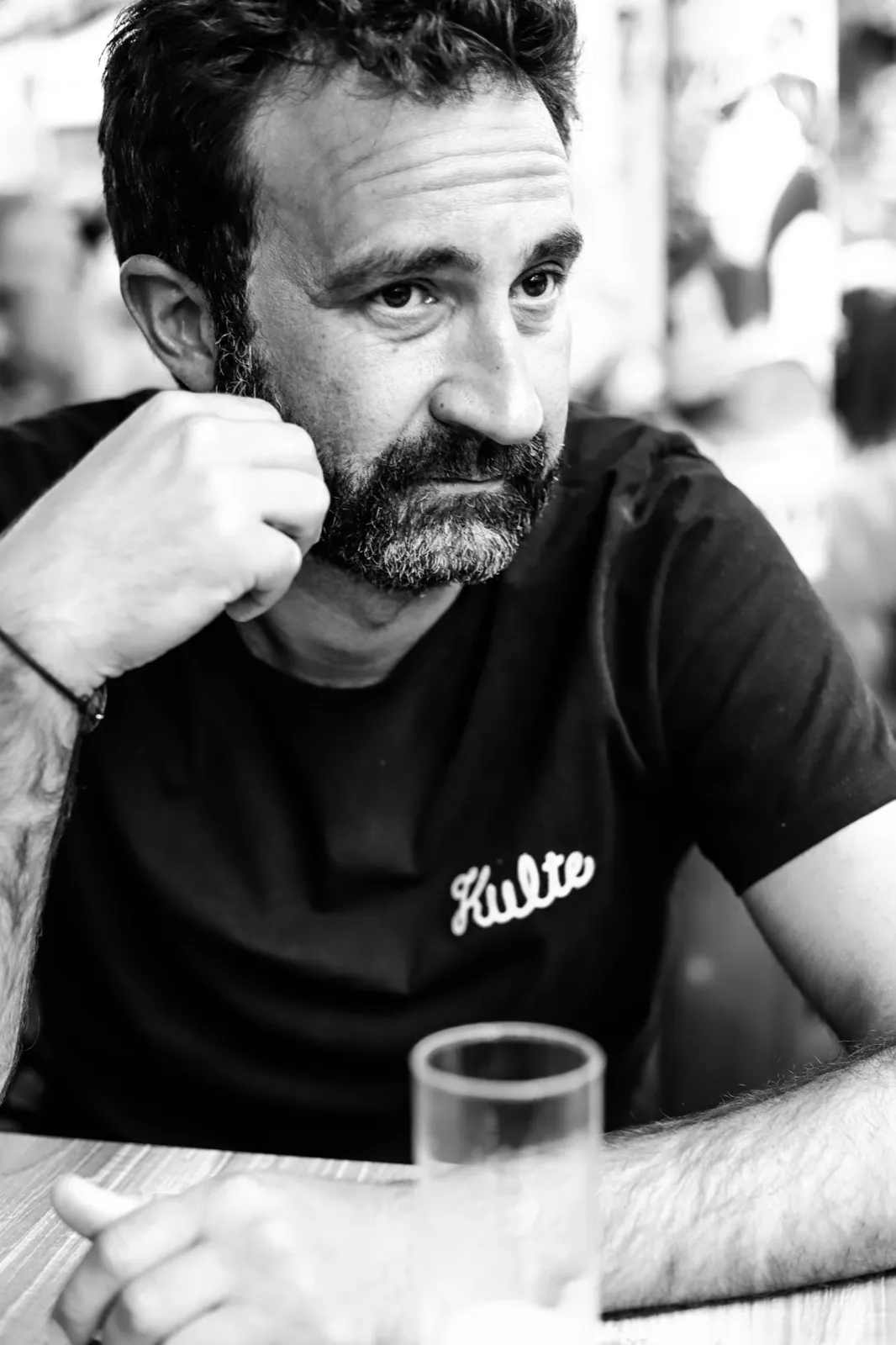 Portrait en noir et blanc d’un homme barbu appuyé sur sa main, assis à une table avec un verre d’eau.