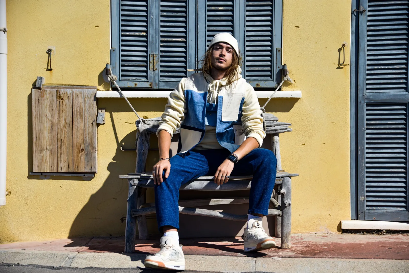 Homme assis sur un banc devant une façade jaune avec volets bleus, habillé de bleu et beige, dans une ambiance estivale.