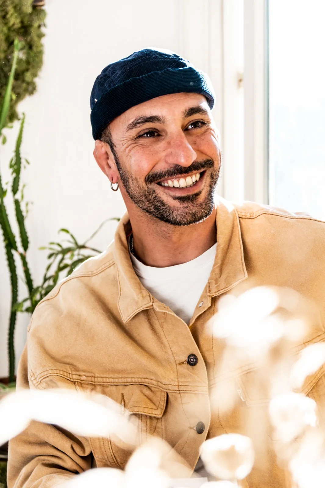 Homme souriant vêtu d’une chemise beige, entouré de fleurs en avant-plan, ambiance naturelle et lumineuse.
