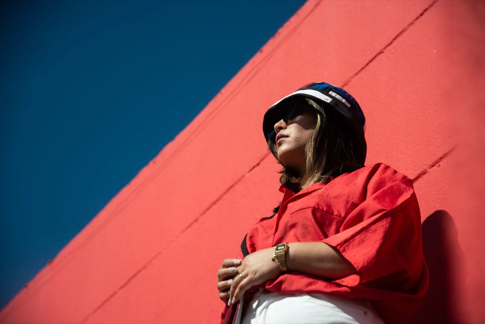 Femme en chemise rouge appuyée contre un mur rouge vif, casquette noire et regard tourné vers le lointain.
