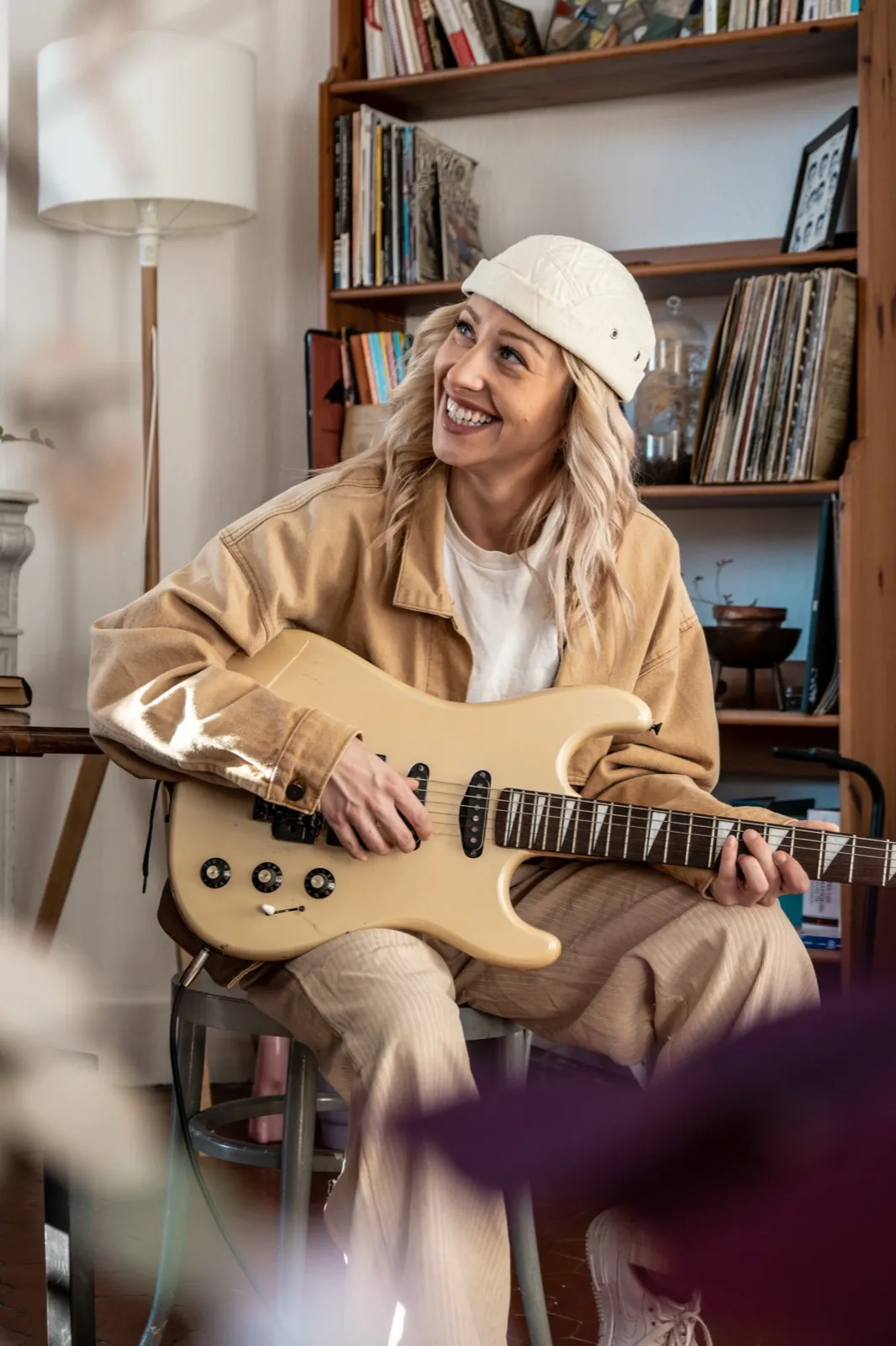 Femme assise dans un salon, souriante, tenant un oud, entourée de livres et de lumière chaleureuse.