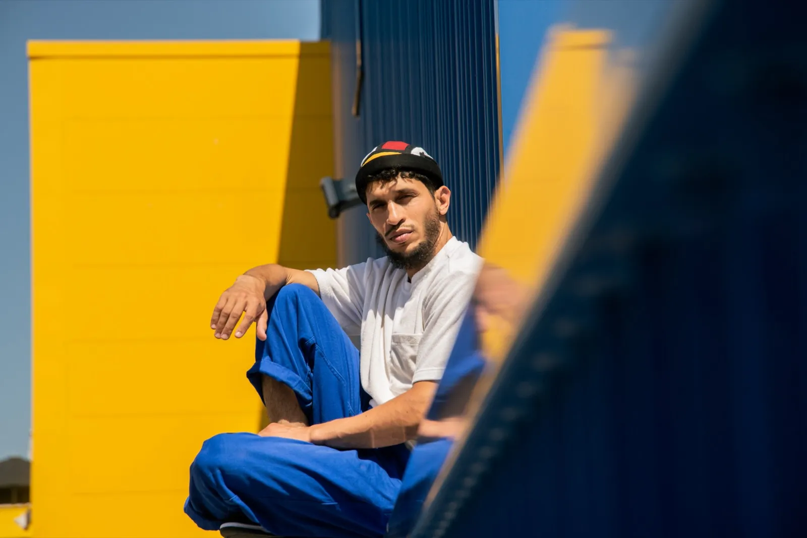 Homme assis sur des marches entre un mur jaune et une rambarde bleue, habillé de blanc et bleu, lumière vive de midi.
