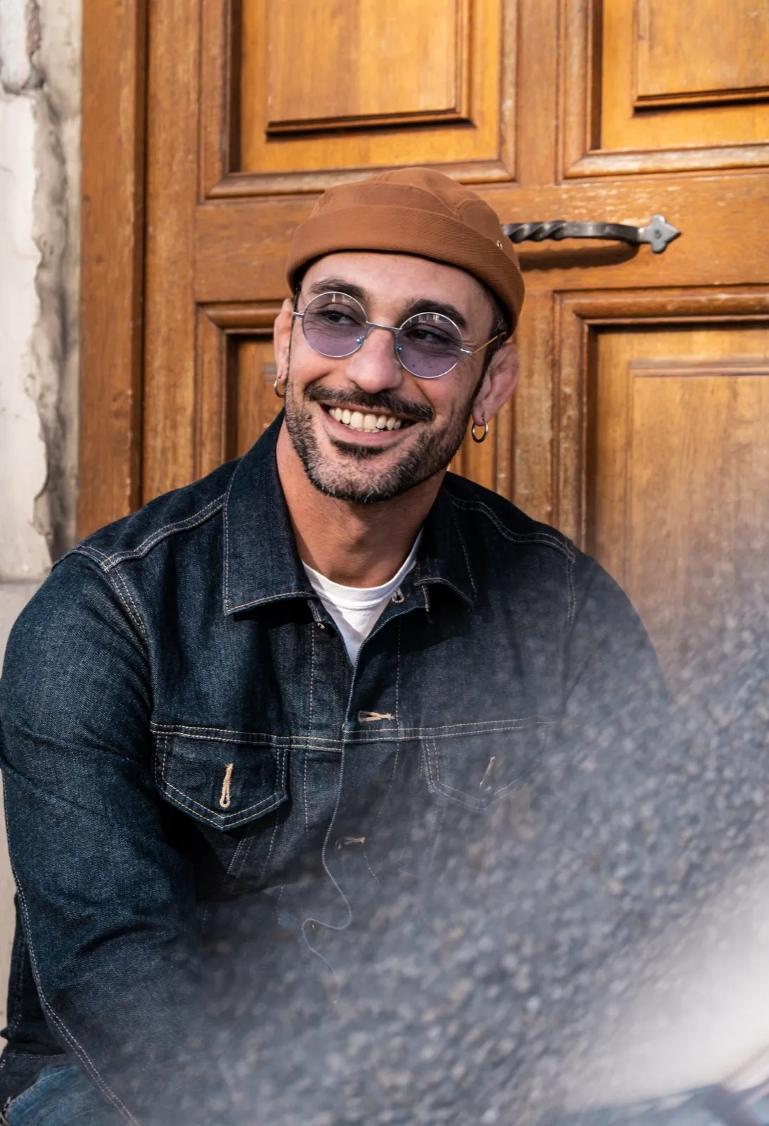 Homme souriant devant une porte en bois, portant une casquette et une veste en jean, lumière douce.