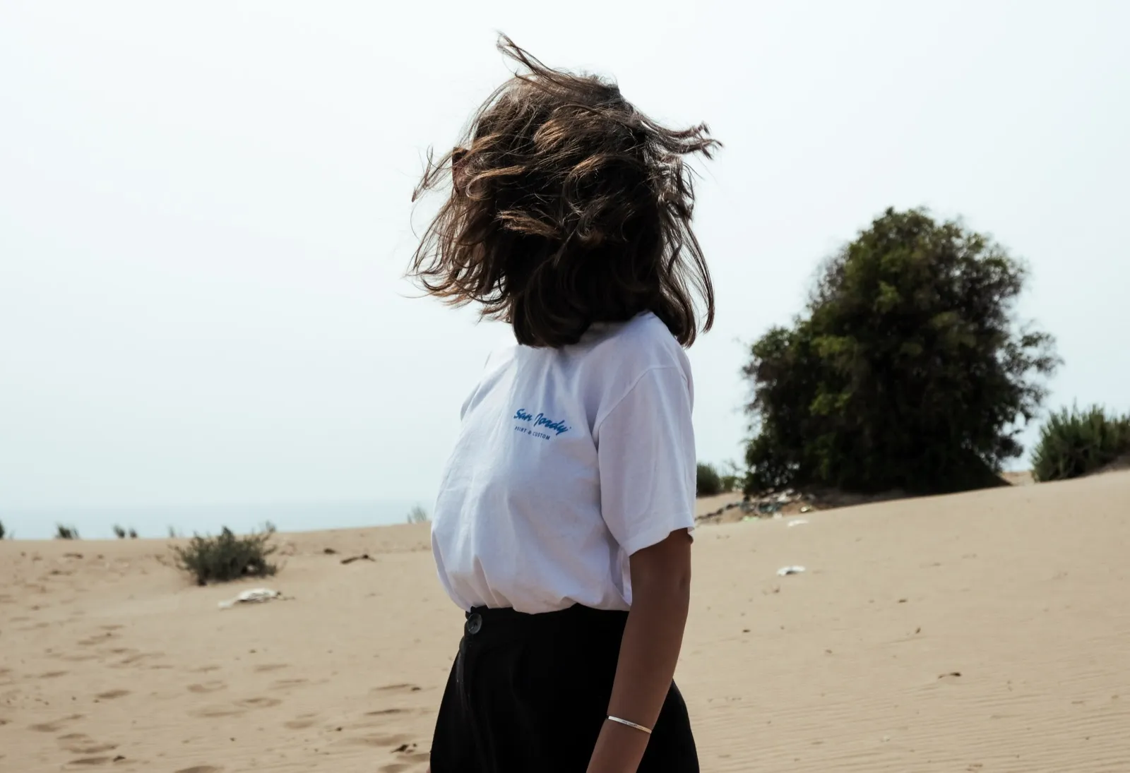 Jeune femme de dos sur une plage, cheveux au vent, face à l’horizon.