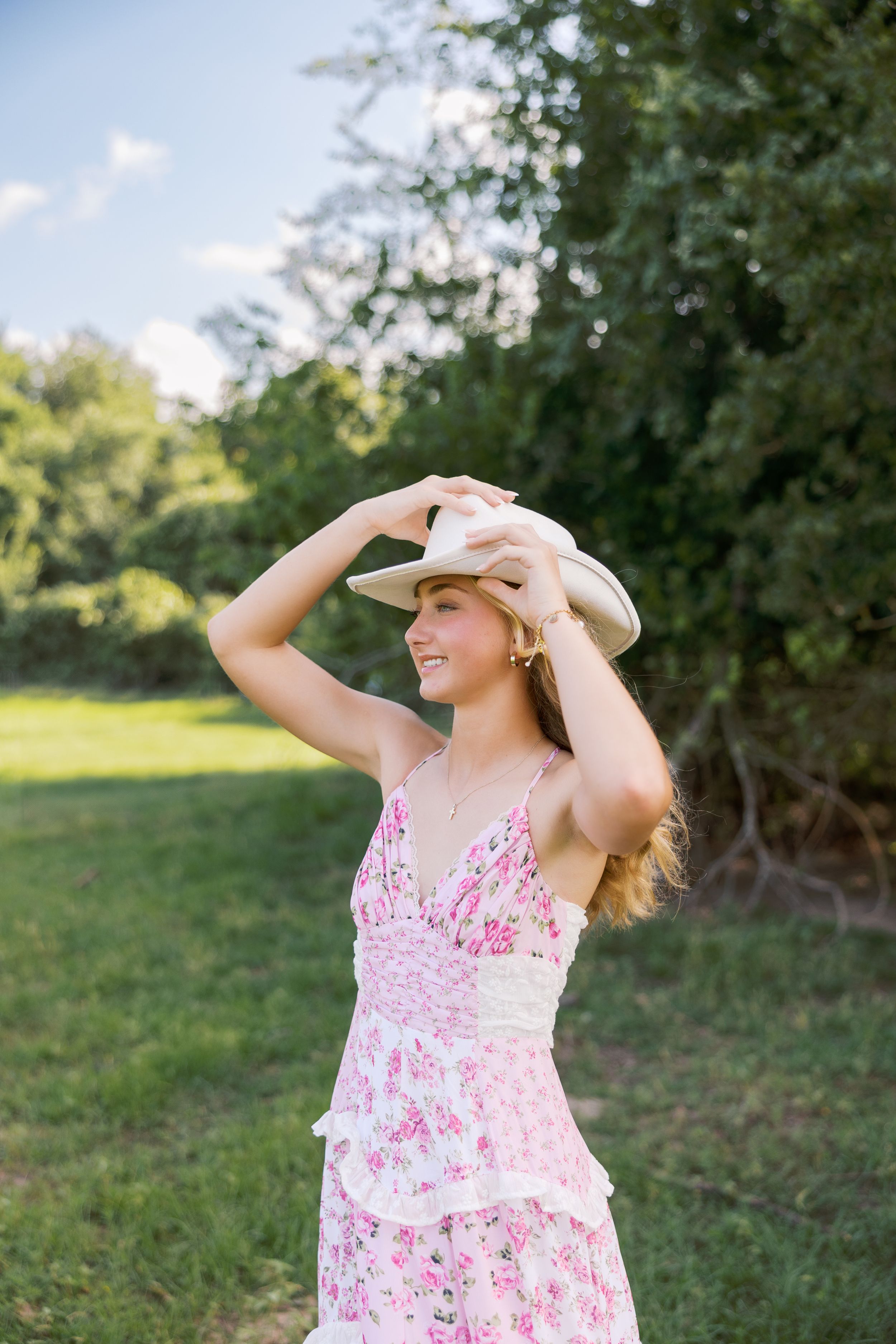 Woman in floral dress twirling outdoors while holding sun hat in summer garden setting.