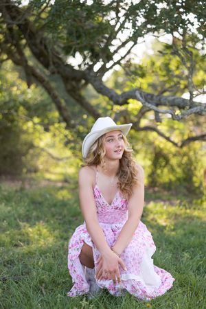 A dreamy outdoor portrait in a pink floral dress and white cowboy hat against greenery.