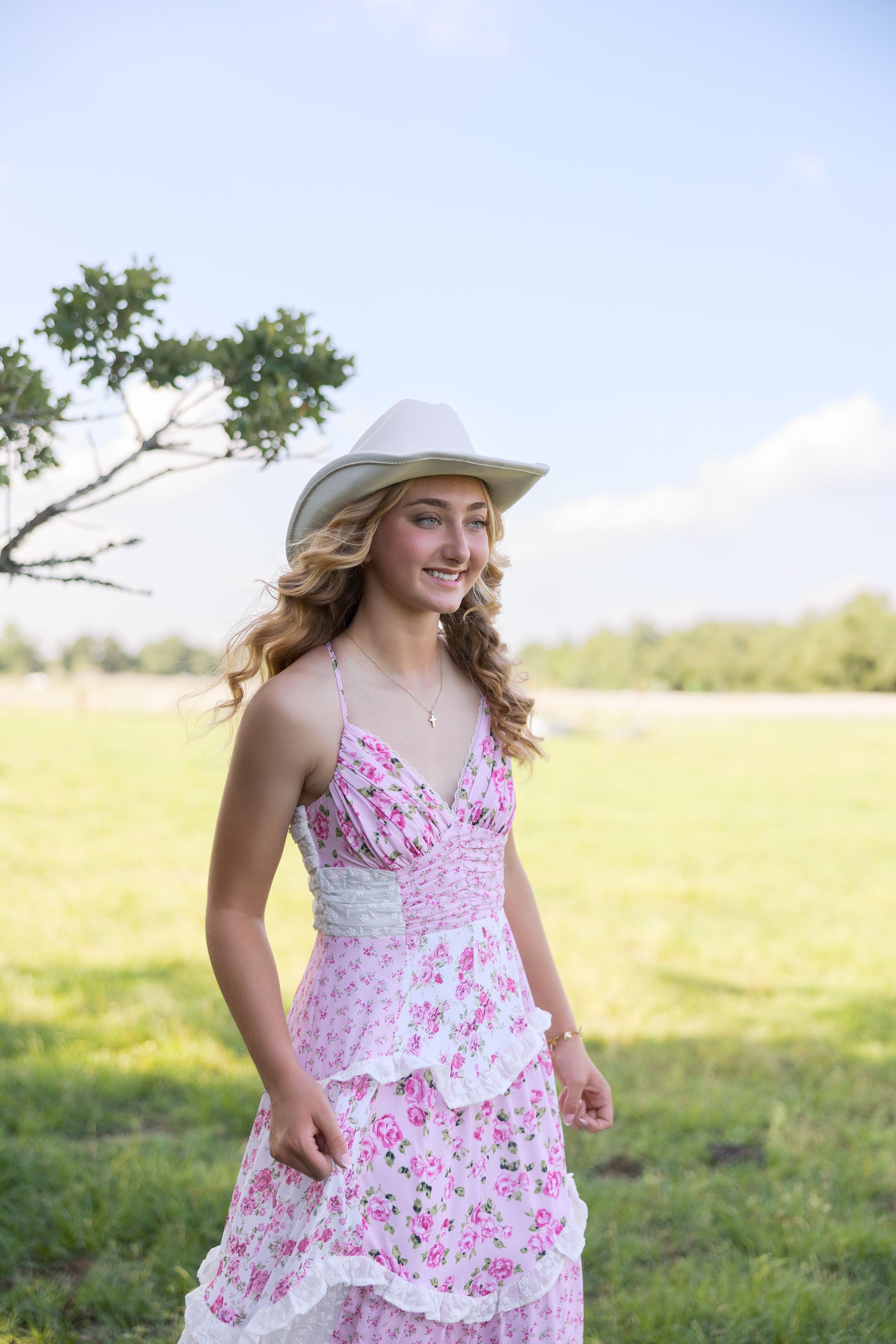 Woman in pink floral dress and sun hat poses elegantly in sunny meadow.