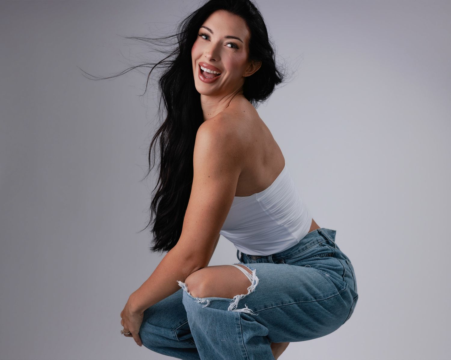 A person in a white t-shirt and distressed jeans sits barefoot against a light studio background, showing red nail polish.