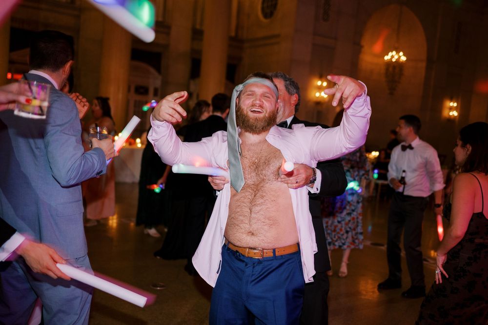 Shirtless guest dances enthusiastically with arms raised at evening reception wearing an open white blazer at Hall of Springs in Saratoga.