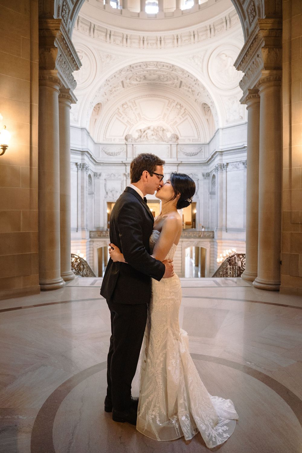 Wedding couple shares intimate moments in an elegant marble hall with domed ceiling and classical architecture.