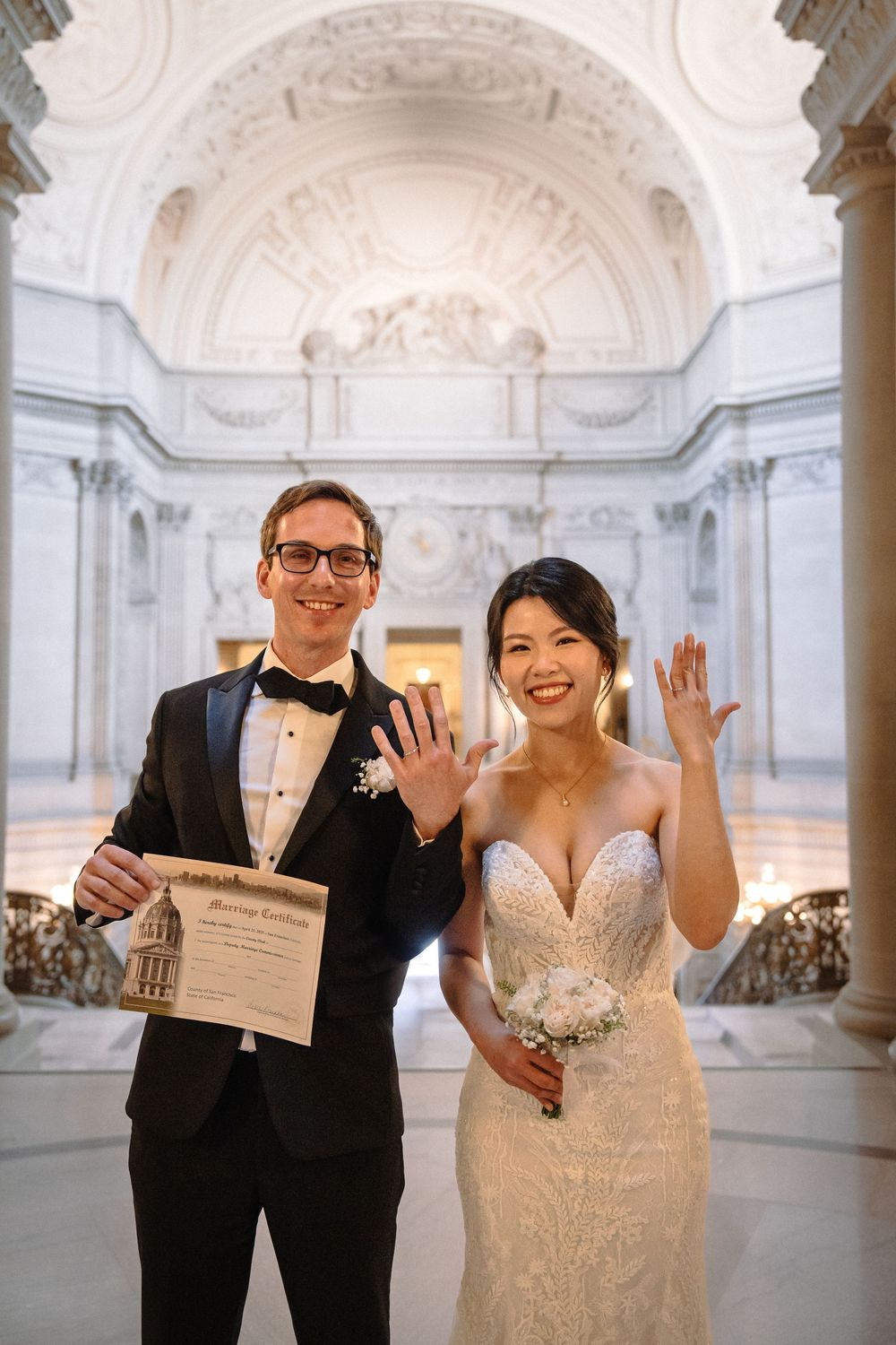 Newlyweds celebrate with joyful expressions while holding their marriage certificate in a marble hall.