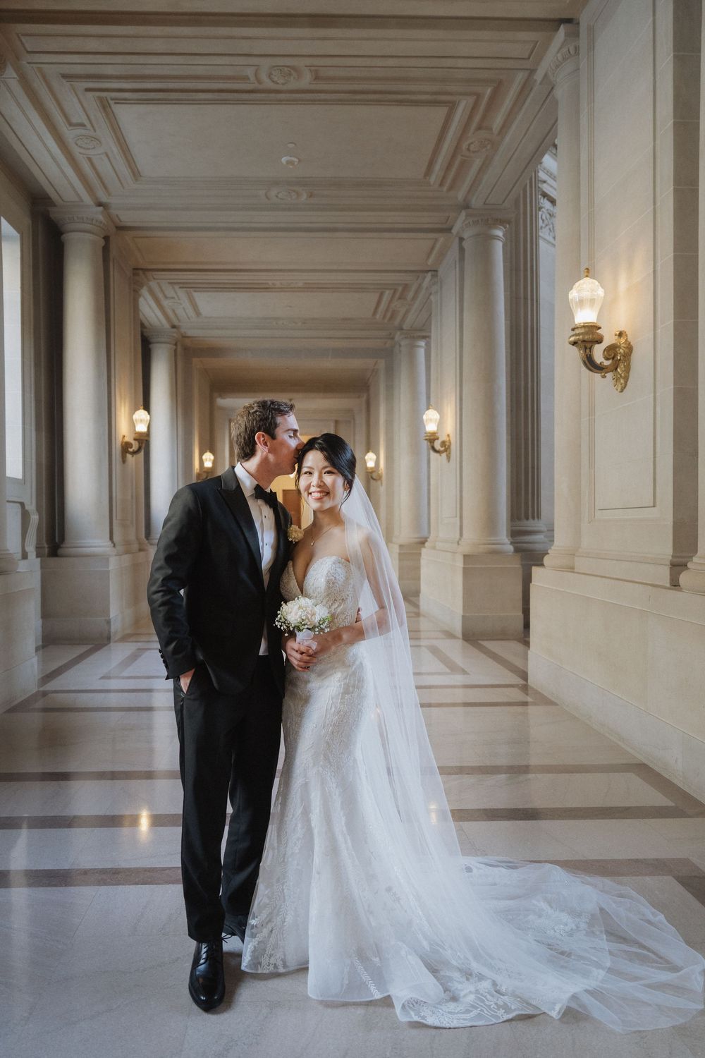 Wedding couple poses in a grand hallway with marble columns and wall sconces.