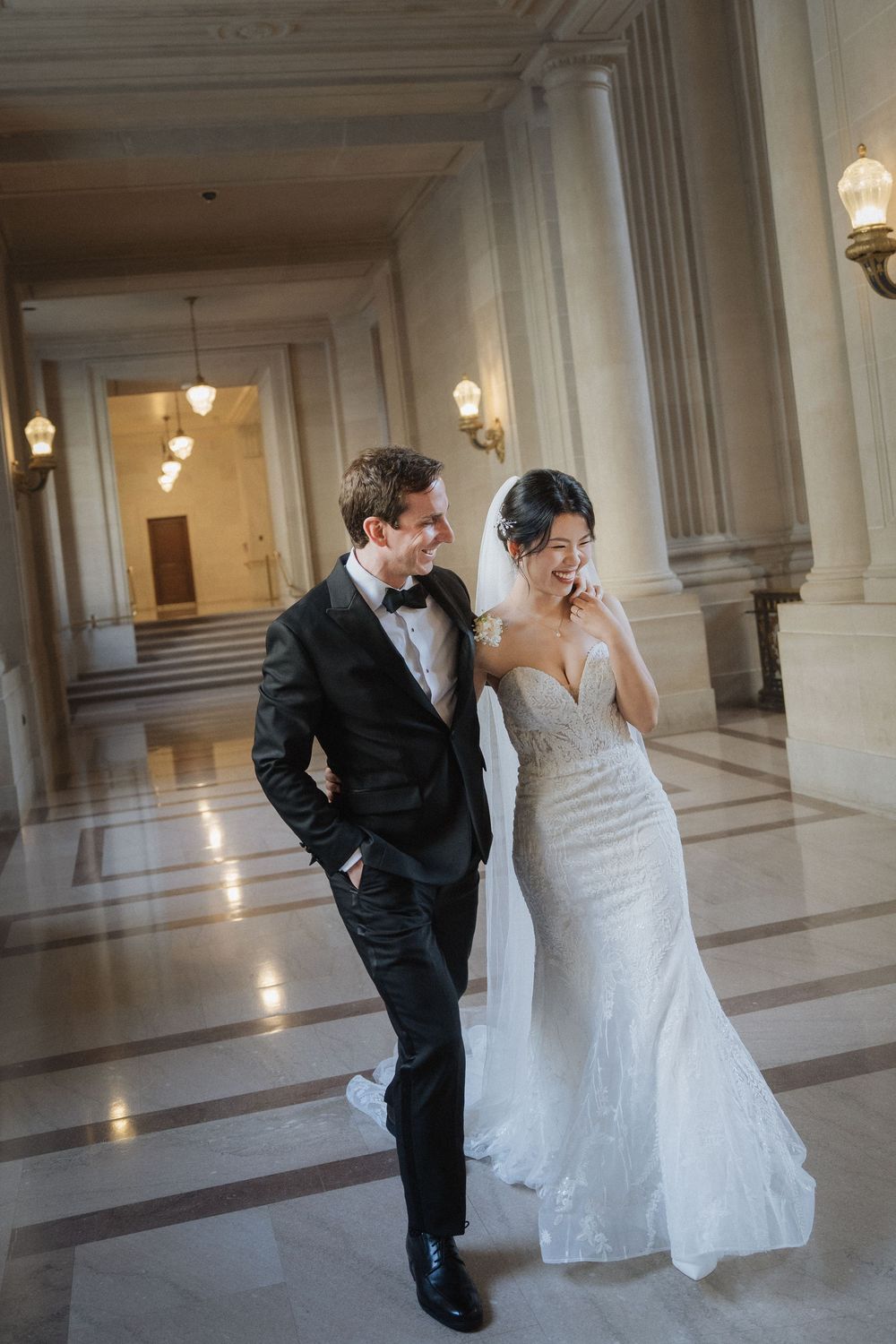 Candid wedding moment in elegant marble corridor with classical columns and pendant lighting.