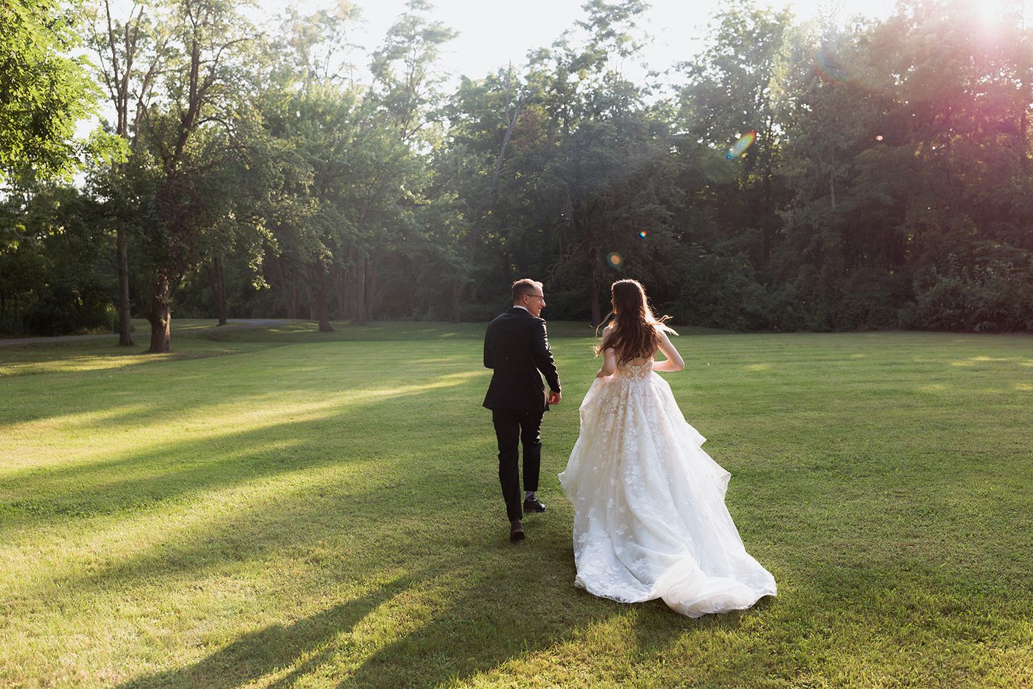 Couple walks hand in hand across a sunlit meadow during sunset.