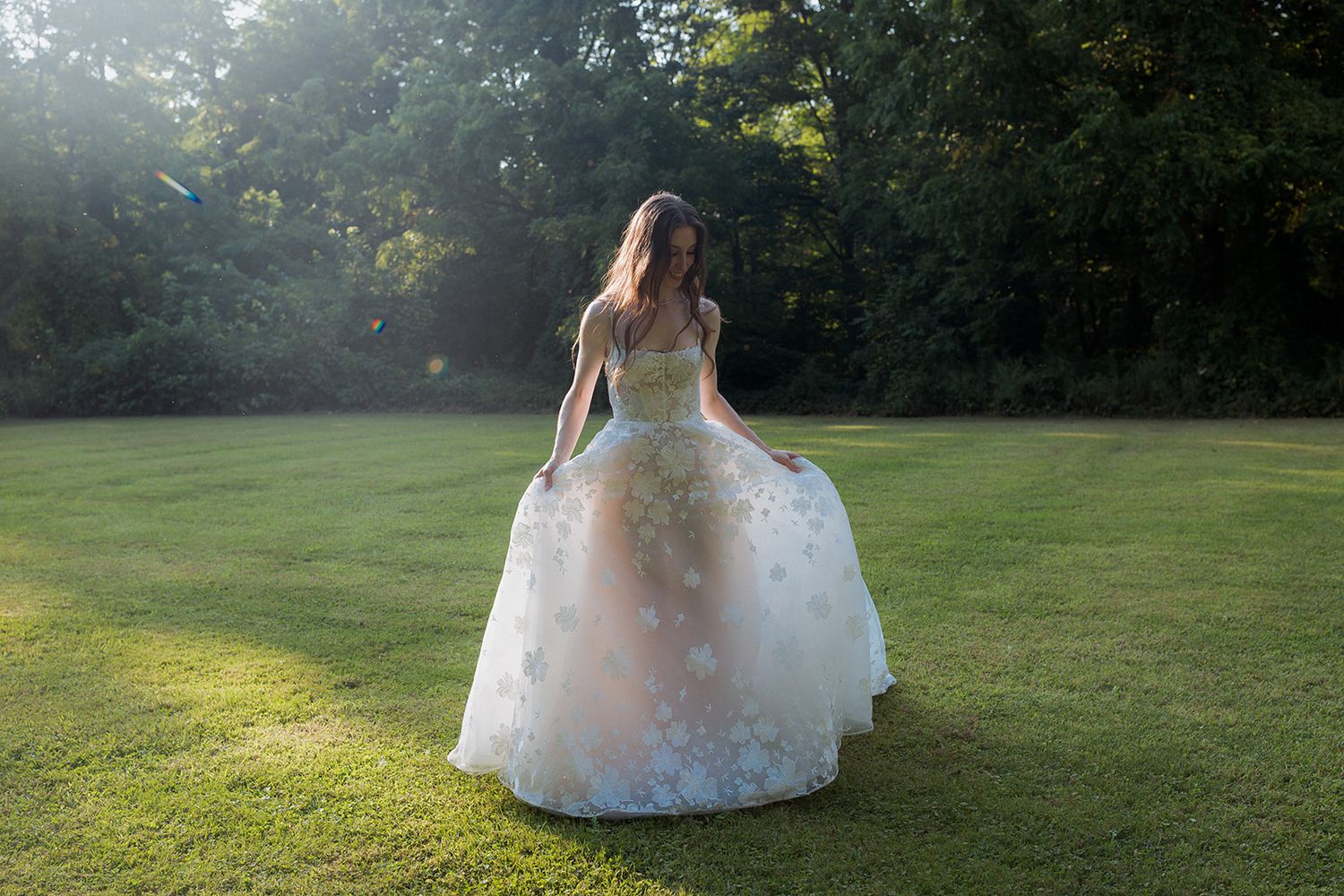 Bride in flowing white gown walks across sunlit lawn during golden hour.