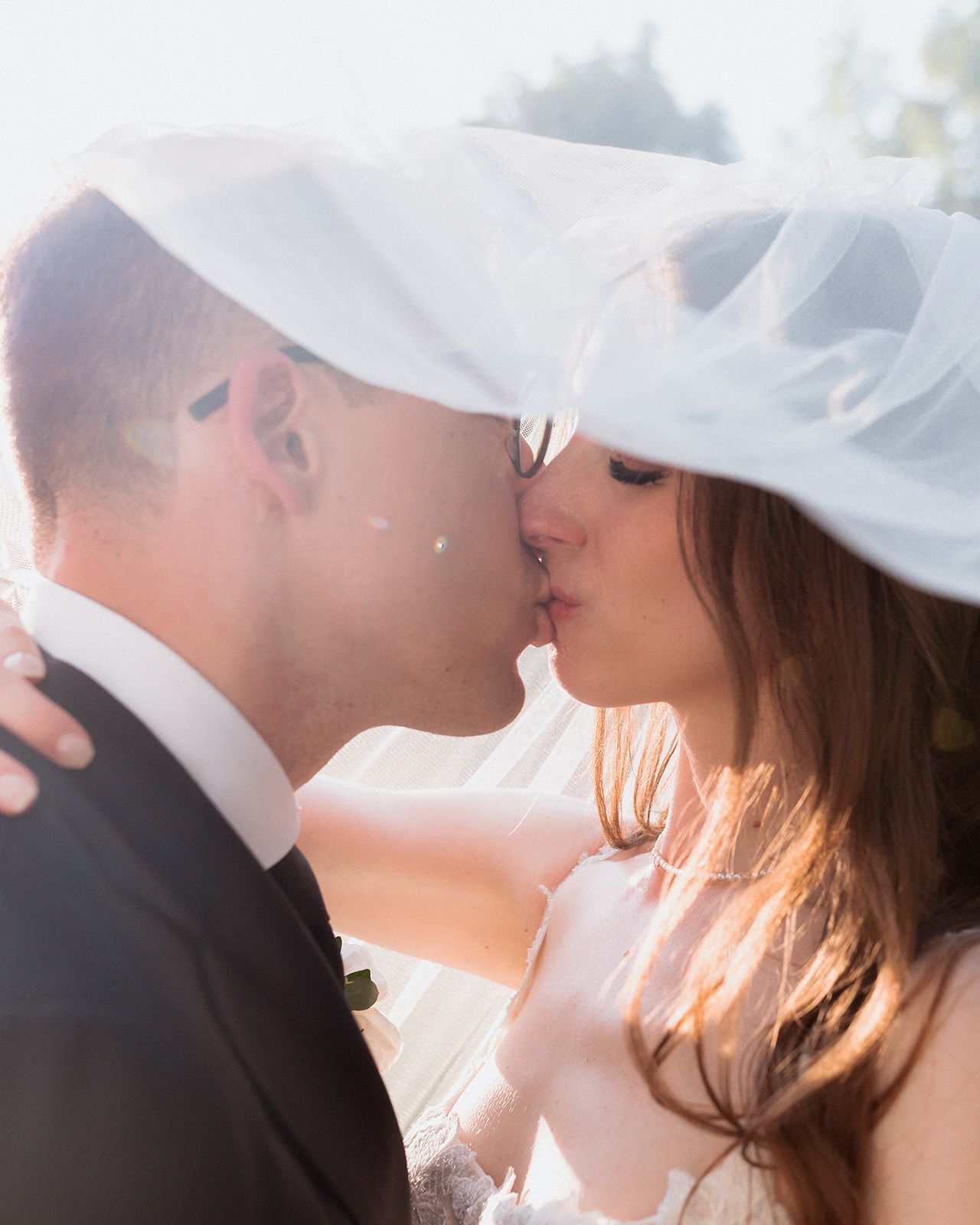 Intimate kiss between couple under a white veil with sunlight streaming through.
