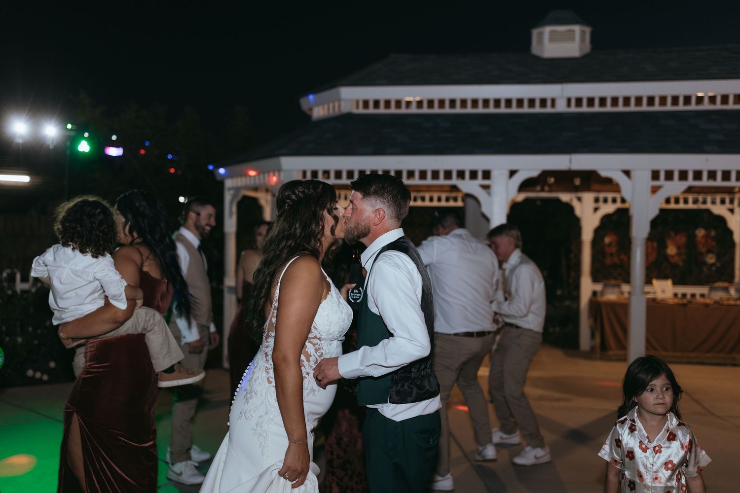 Couple in wedding attire sharing a kiss on a dance floor at an outdoor reception at storybook garden in hanford, ca, with guests dancing too