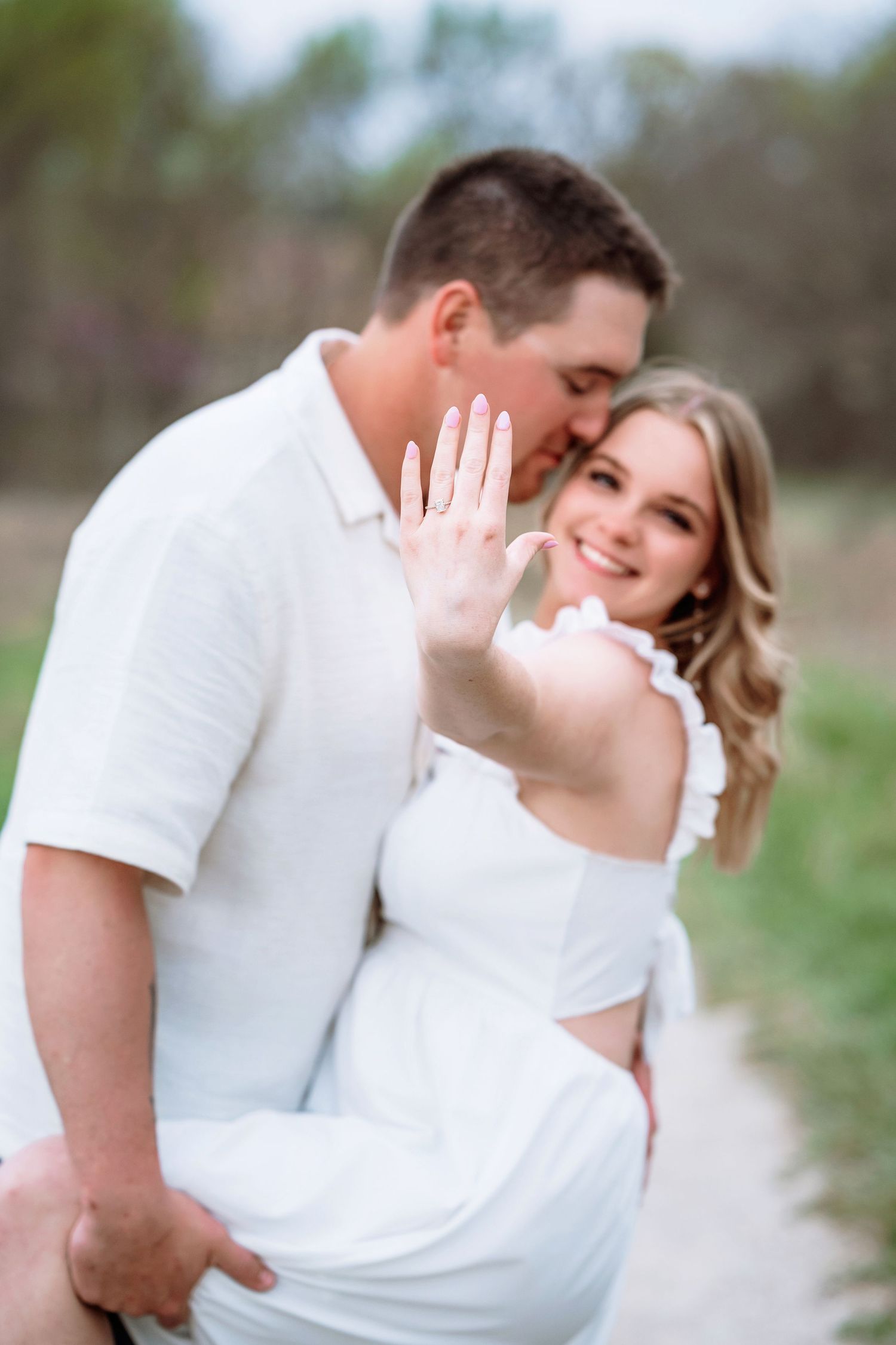 A romantic engagement photo showing a couple in white outfits embracing outdoors while showing off a ring.