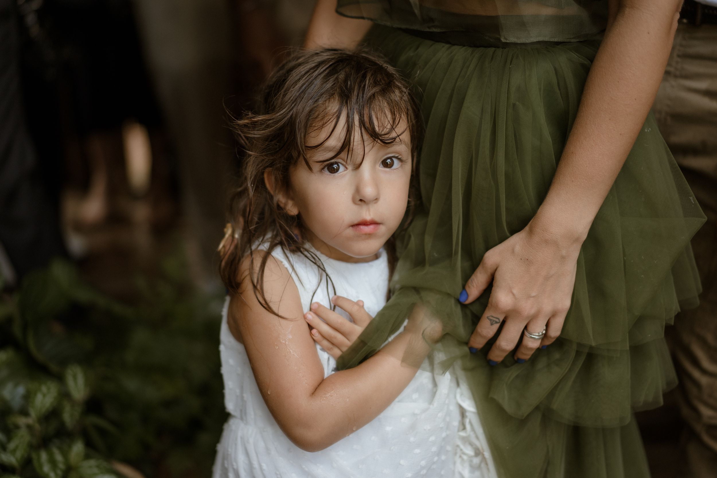 Young attendee in a white dress stands during an outdoor wedding celebration.