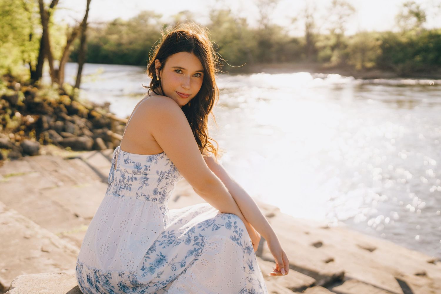 A woman in a flowy white and blue sundress sits at the edge of a sunlit river during golden hour.