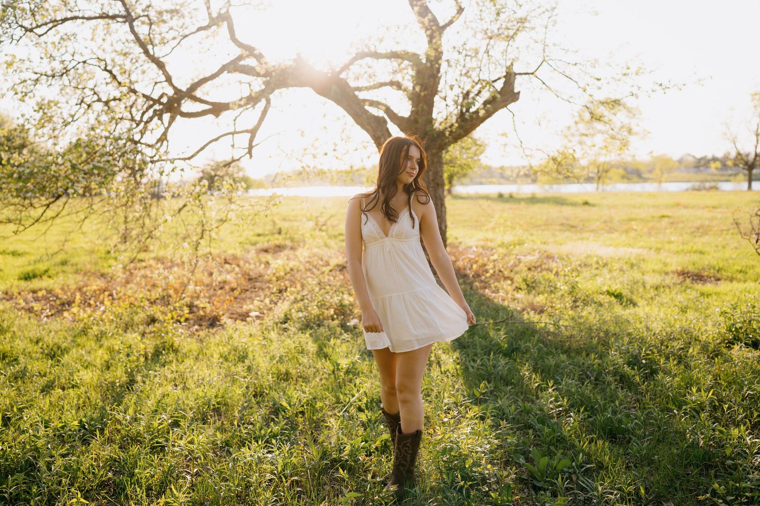 Someone in a white sundress and boots stands under a sunlit tree in a pastoral field at golden hour.