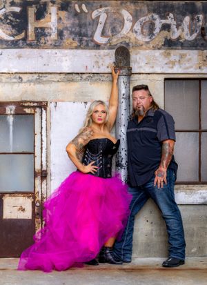 Couple poses against weathered industrial wall with vintage signage.