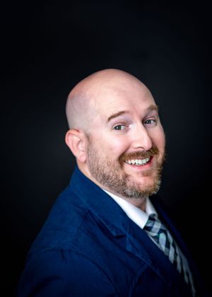 Professional headshot of bald man with beard wearing navy suit and striped tie against dark background.