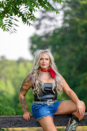 Someone in denim shorts and bandana sitting on wooden fence with trees in background.