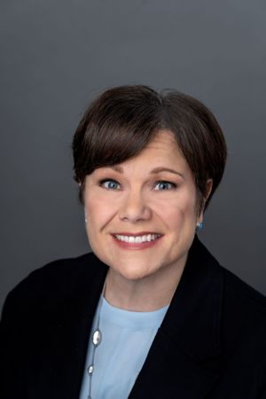 Professional portrait of person in black blazer and blue blouse against grey backdrop.