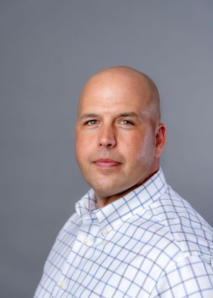 Professional headshot against grey background wearing blue checkered button-down shirt.