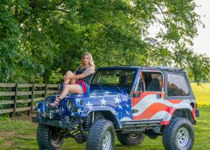 Patriotic themed Jeep with American flag paint job parked under green trees.