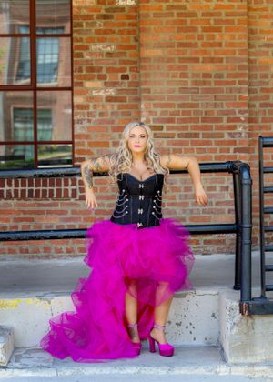 Woman in black corset and pink tulle skirt poses against brick wall.
