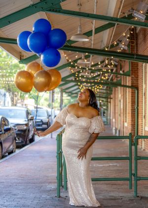 Celebratory photo with gold and blue balloons on a covered brick walkway with string lights overhead.