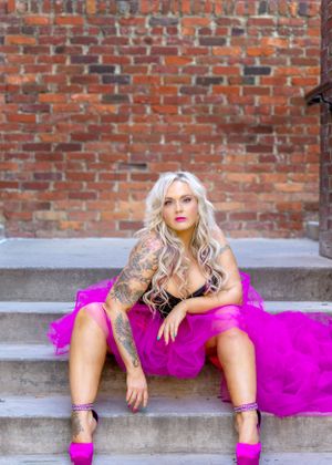 Woman in black corset and pink tulle skirt poses on concrete steps with brick wall backdrop.