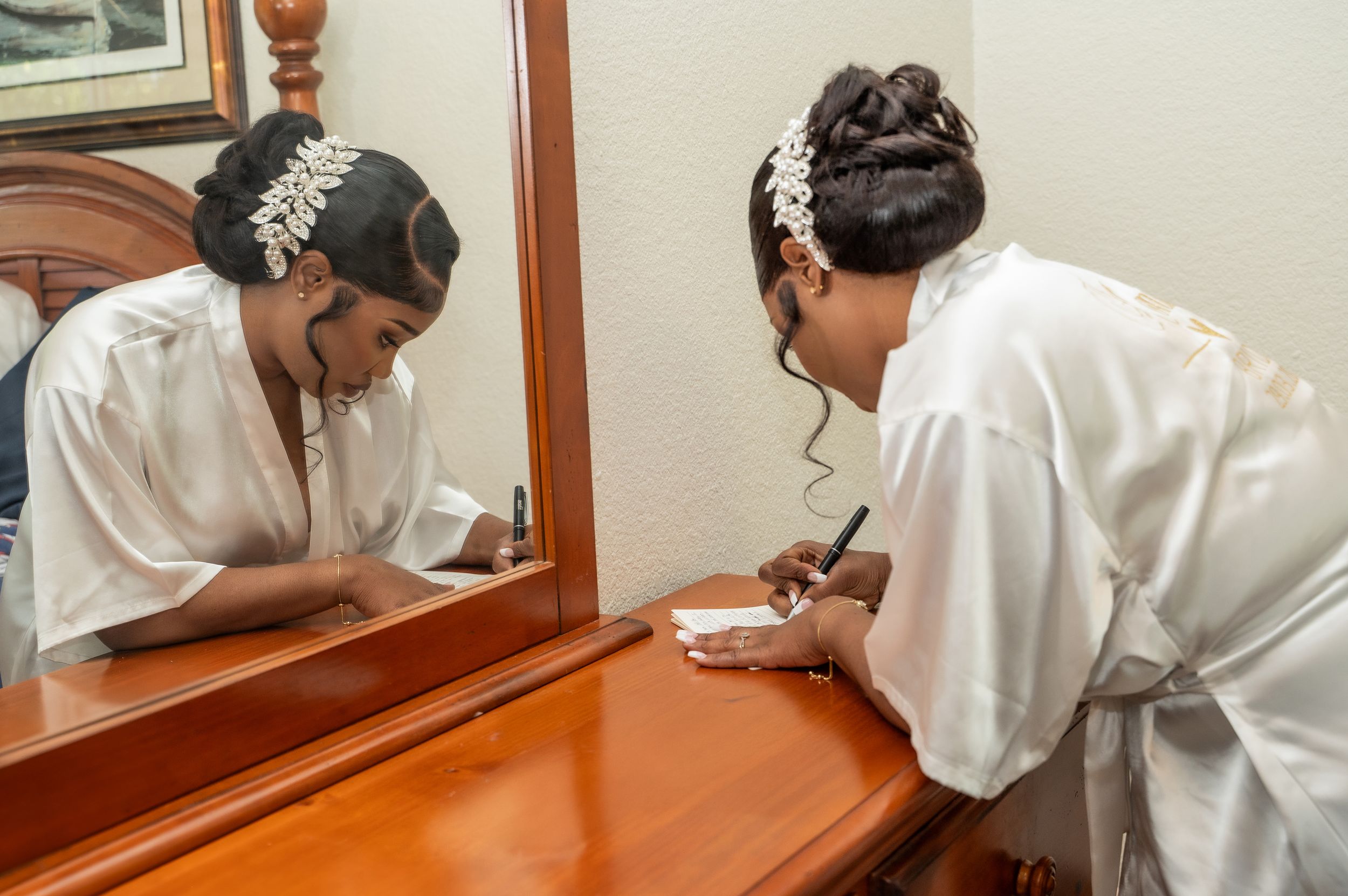 Bride in white robe writing a note by wooden dresser mirror, wearing elegant hair accessory on wedding day.