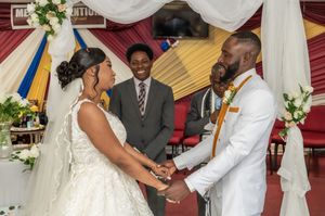 A couple exchanges vows at a wedding ceremony, holding hands before an officiant in a decorated church.