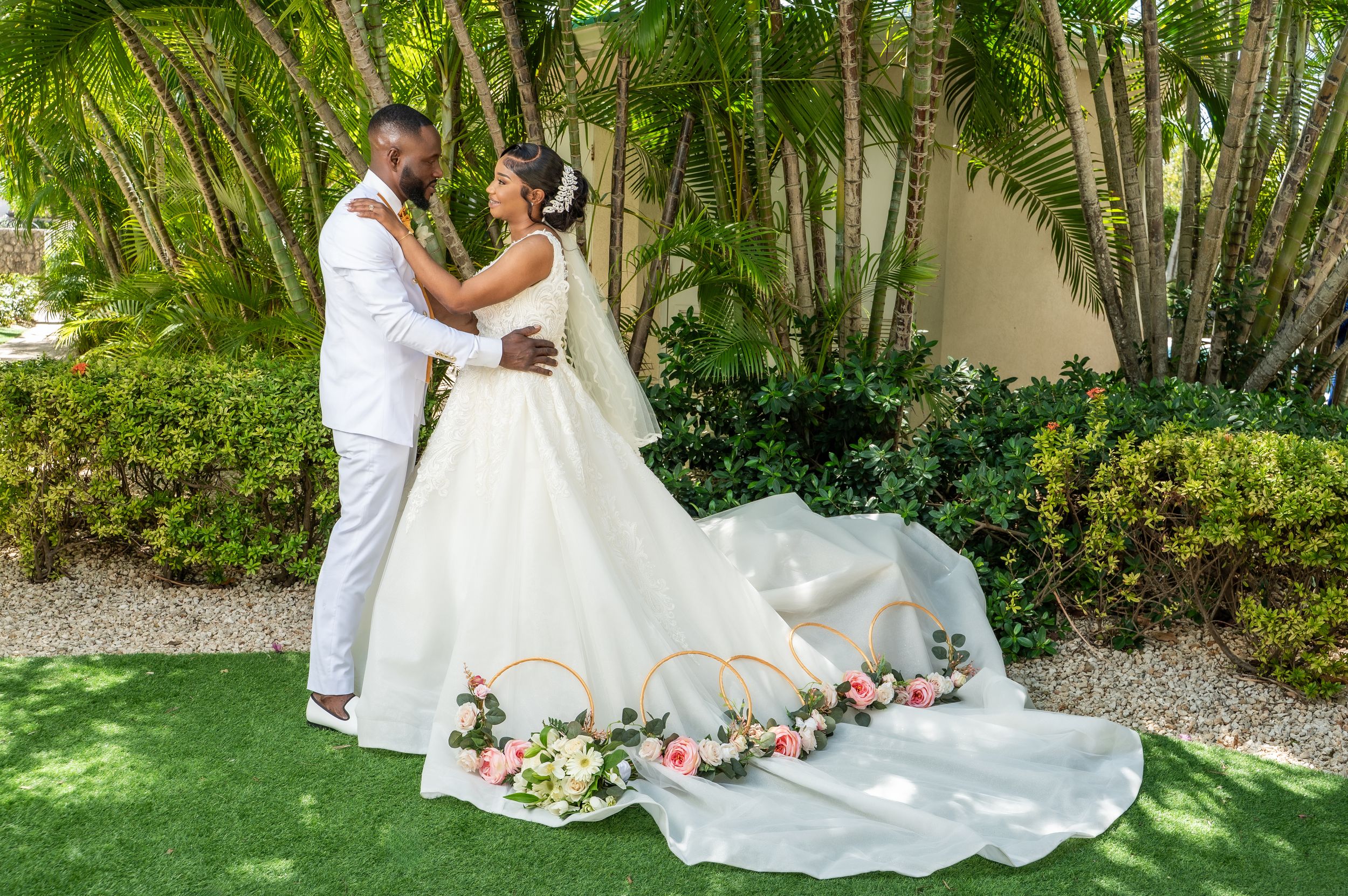 A bride and groom embrace outdoors on green grass, surrounded by tropical palms and floral hoop decorations.