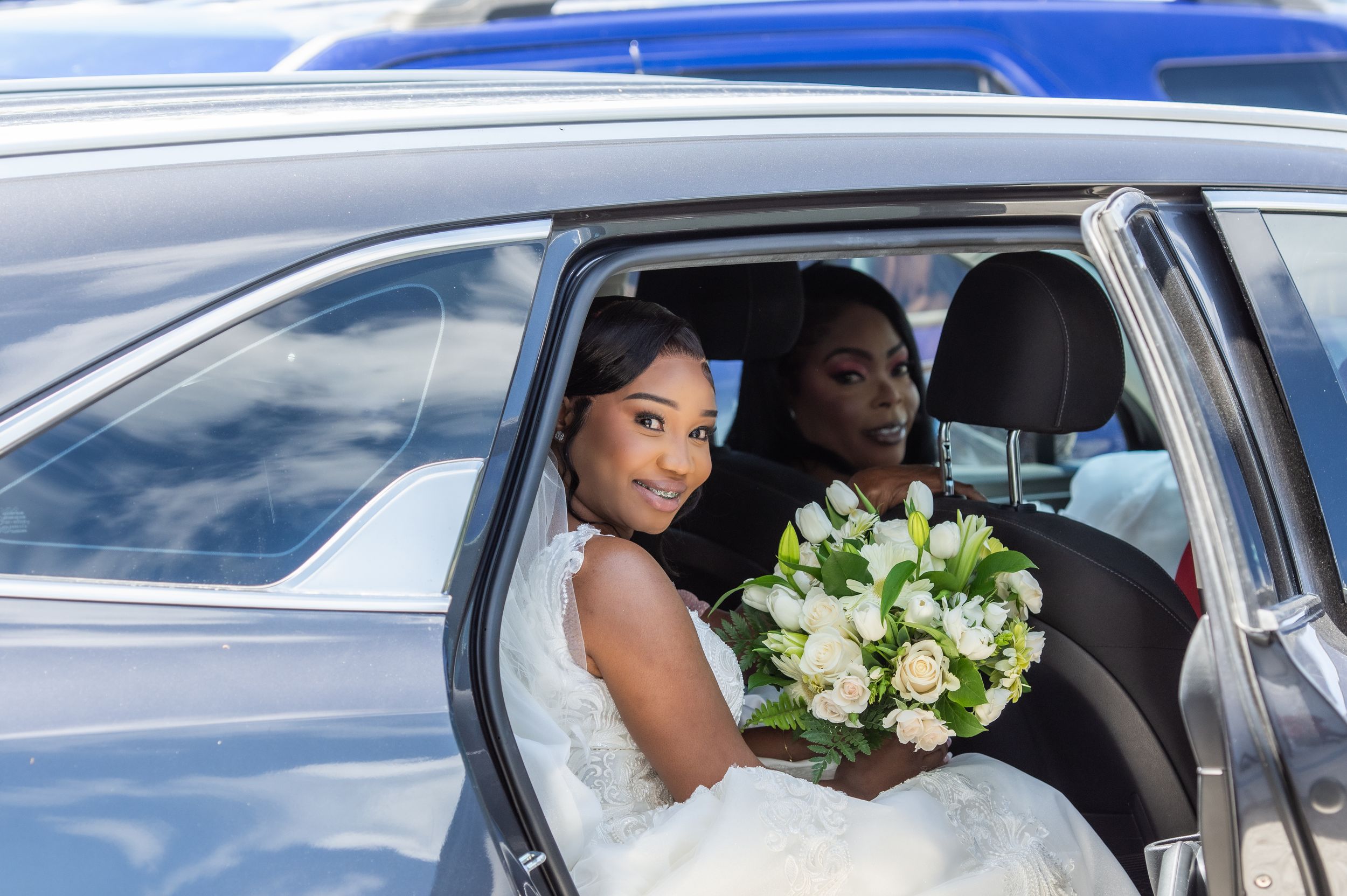 Bride in white wedding dress holding bouquet of white flowers sits in luxury car with guest behind her.