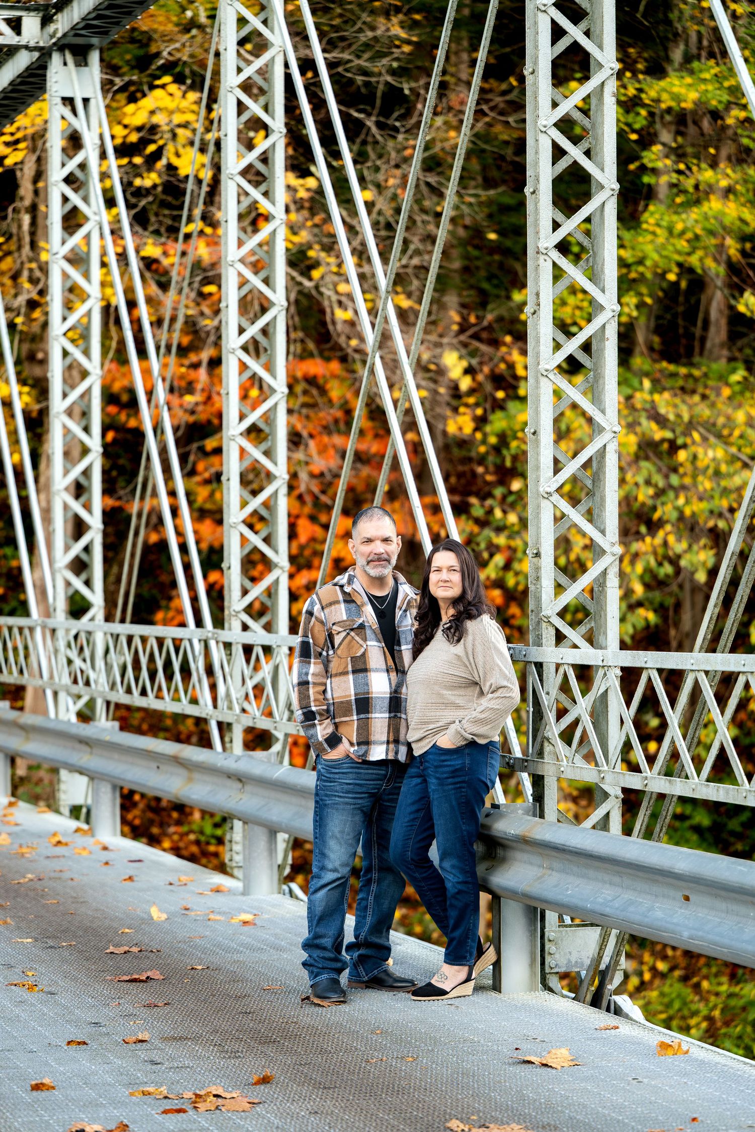 Couple in casual flannel and jeans pose together on a steel truss bridge surrounded by vibrant orange and yellow autumn foliage. youngstown