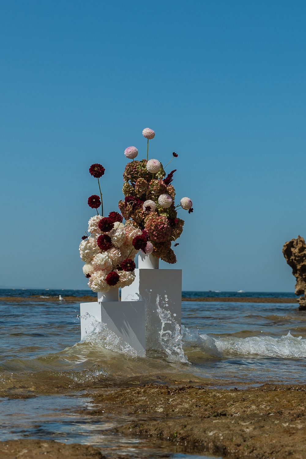 White and burgundy floral arrangements on pedestals getting splashed by ocean waves.
