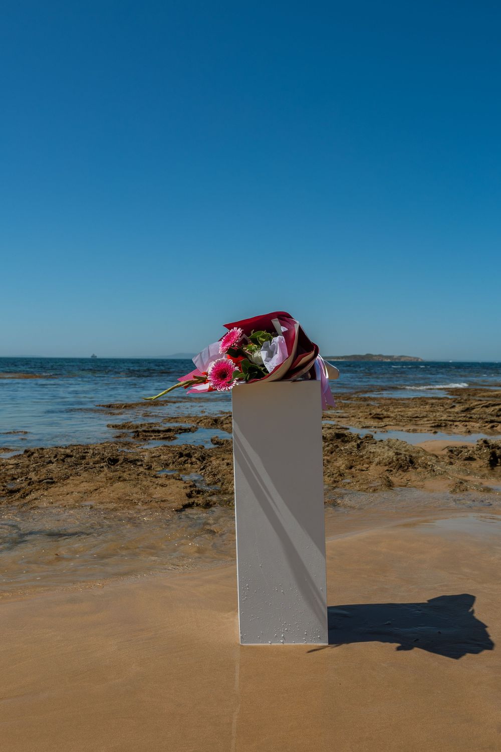 A single white pedestal with pink flowers displayed on a sandy beach.