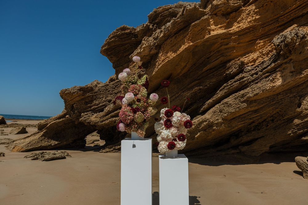 Floral arrangements displayed on white pedestals against a rocky beach backdrop.