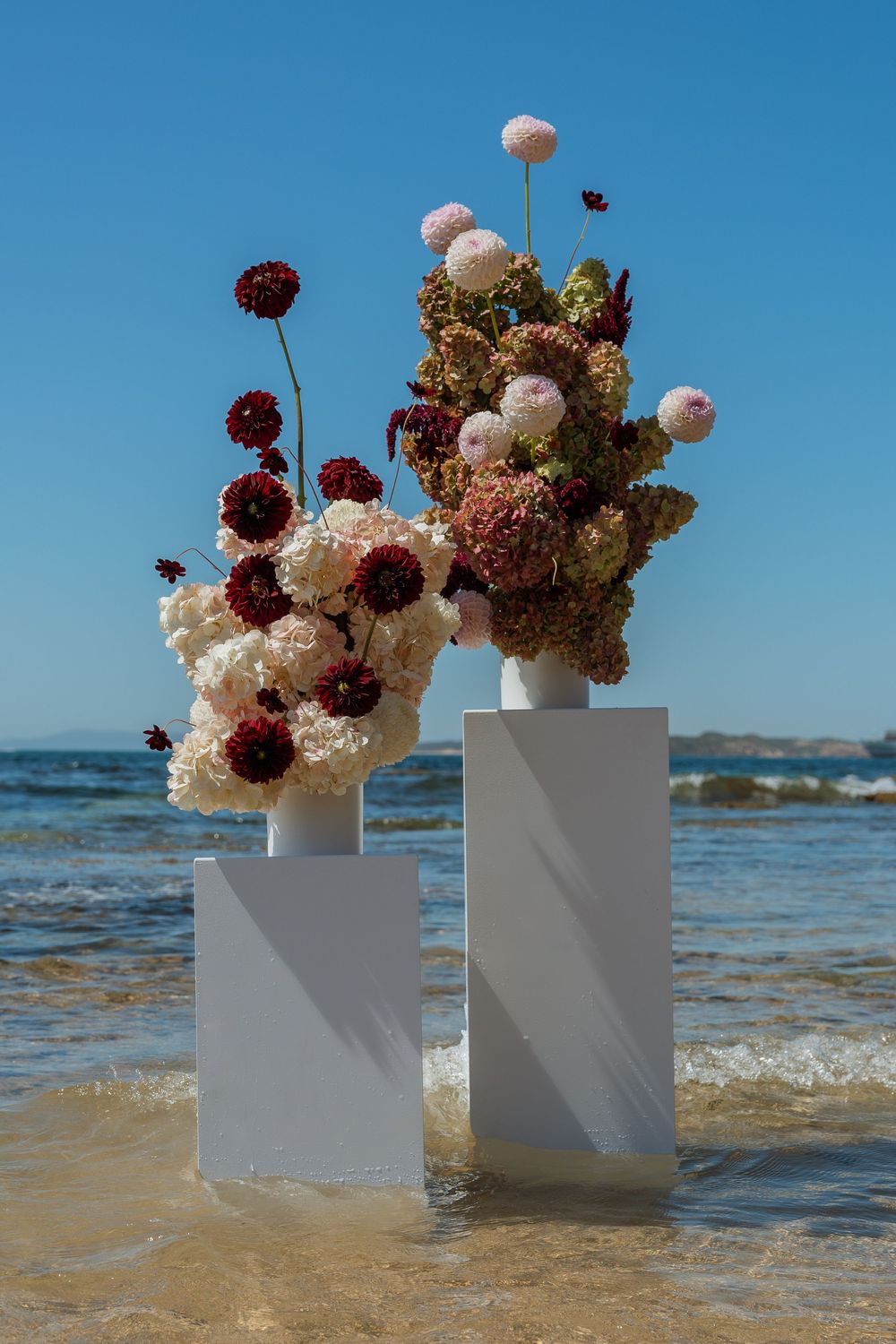 Pink and burgundy flower arrangements on white pedestals with ocean waves in the background.