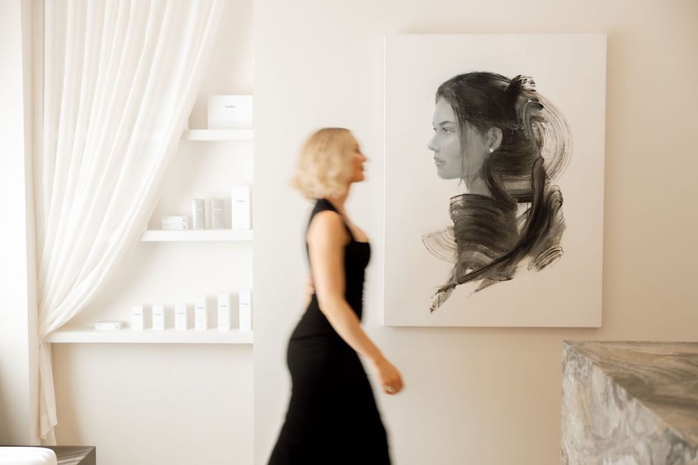 A person in a black dress walking past a modern art portrait in a minimalist white room.