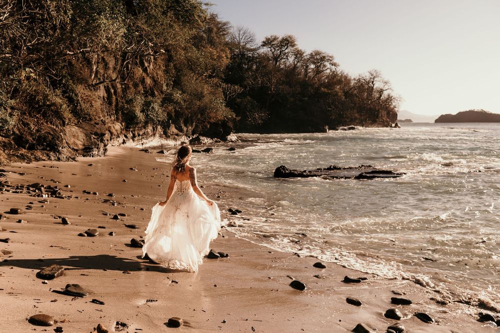A figure in a flowing white dress walks along a scenic rocky beach with waves crashing on the shore.